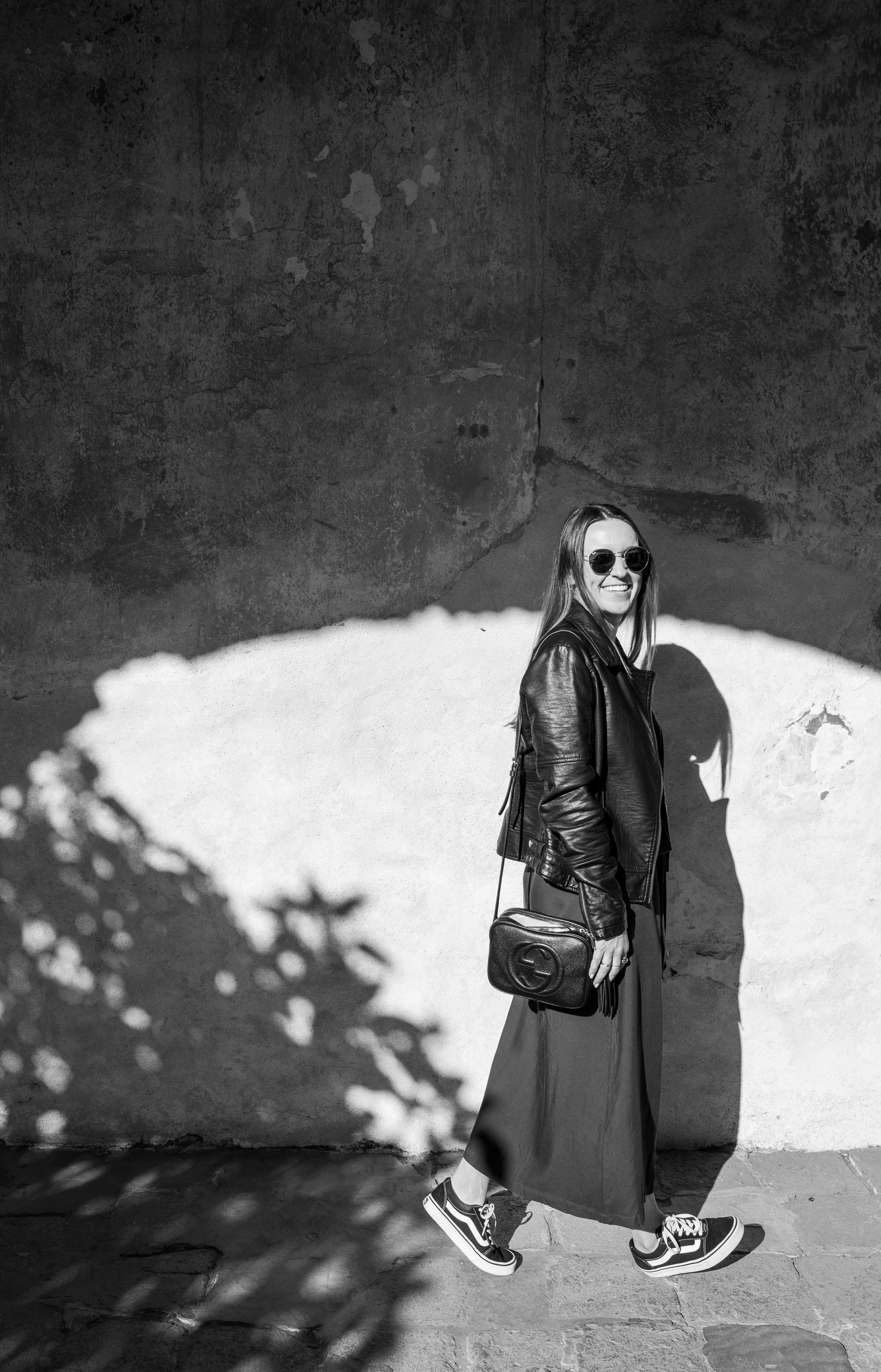 grayscale photography of woman wearing leather zip-up jacket and sunglasses walking on pathway smiling